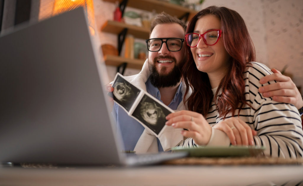 Pareja de hombre y mujer enseñando una ecografía de bebé en videollamada a un portatil despues de conocer la probabilidad de quedar embarazada a la primera